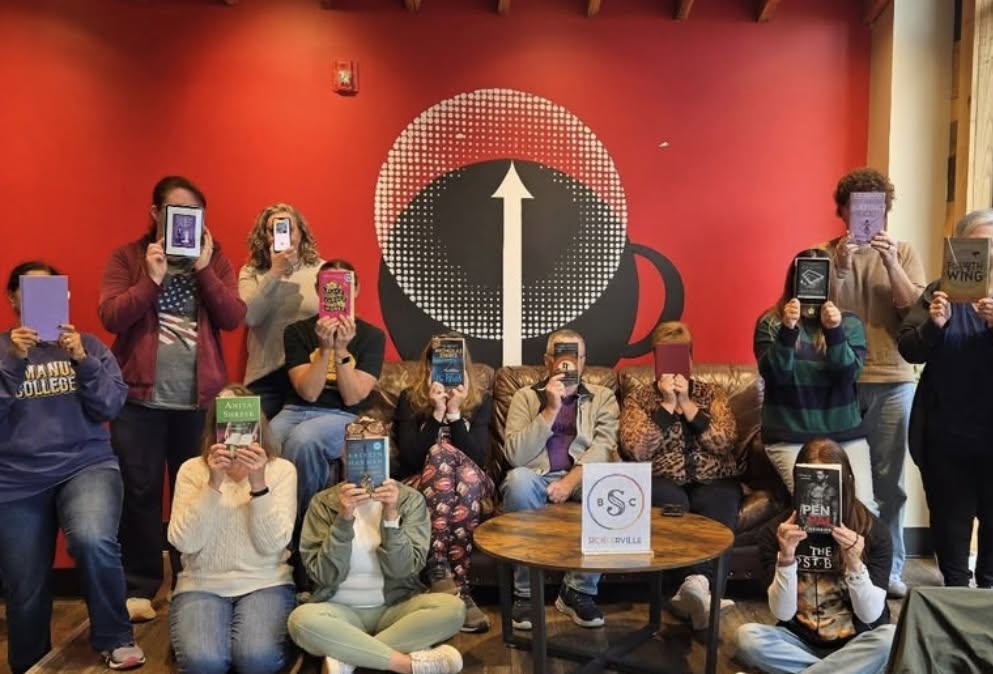 Book club members holding up their books in front of the red coffee cup mural at High Grounds Glassboro